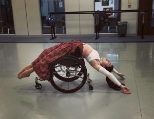 A dancer is bending backwards over a wheelchair. She is wearing a red skit, white top and has long black hair. She is in a dance studio on a white floor with barres behind her.