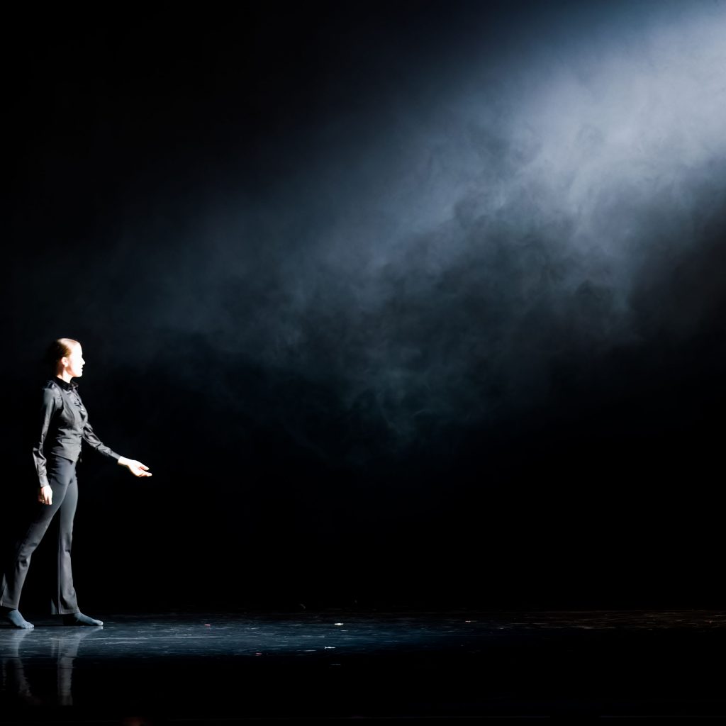 A dancer wearing black is on a dark black stage walking towards a bright white light