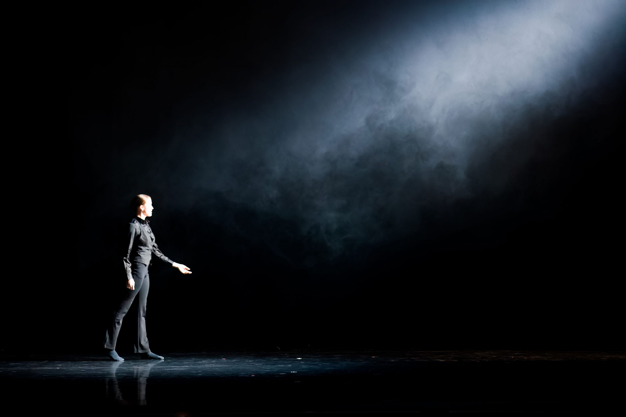 A dancer wearing black is on a dark black stage walking towards a bright white light