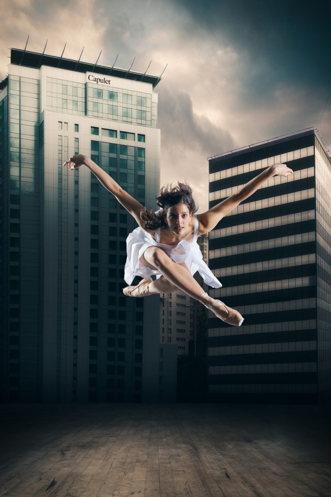 a female dancer with long hair and wearing a white dress is jumping up in the air with her arms outstretched. There are modern buildings and a dramatic cloudy sky behind her.