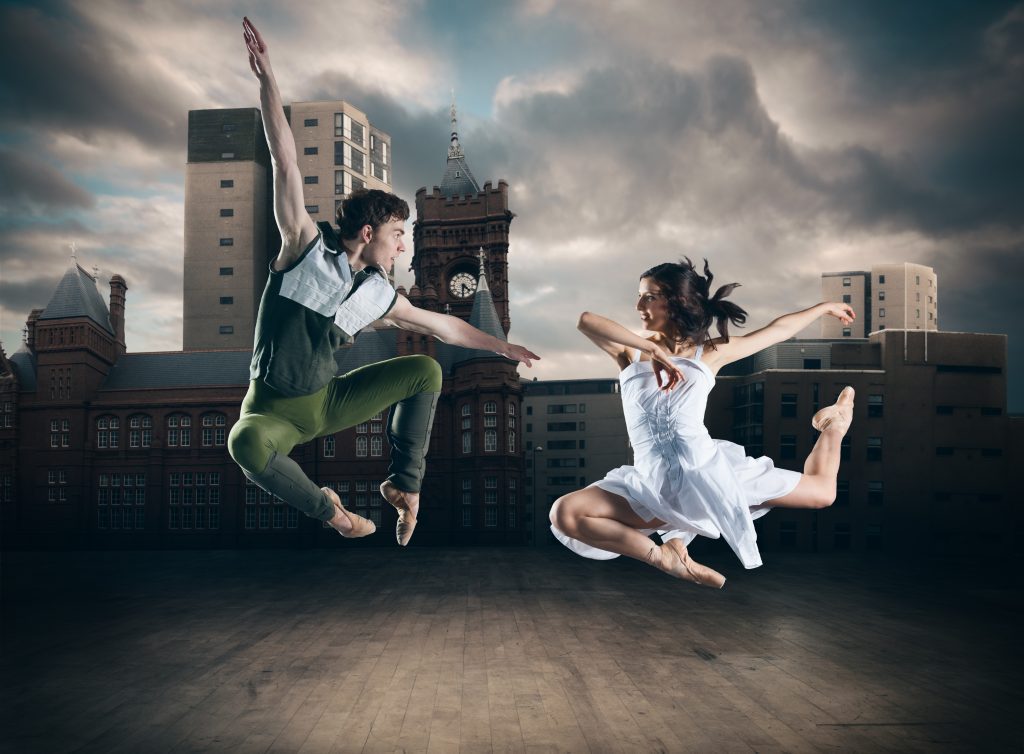 A male and female dancer wearing costumes are jumping in the air and smiling at each other. Behind them are modern buildings and a cloudy sky, and below them is a wooden floor.
