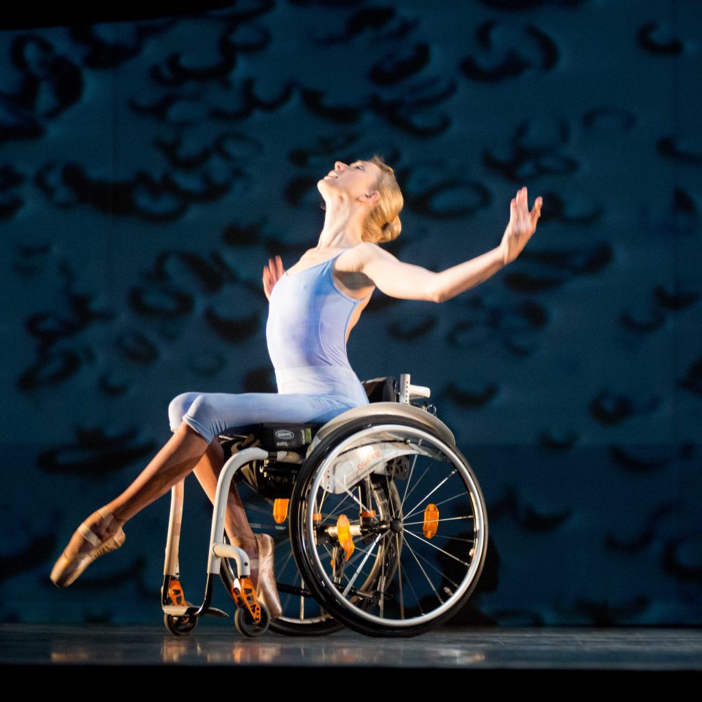 A dancer with short blonde hair and wearing a blue unitard and pointe shoes is sitting in a wheelchair on stage with her arms outstretched and looking upwards. © Sian Trenberth Photography