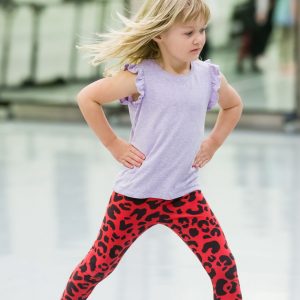 A young girl with blonde hair wearing a white top and red leggings is dancing in bare feet in a dance studio